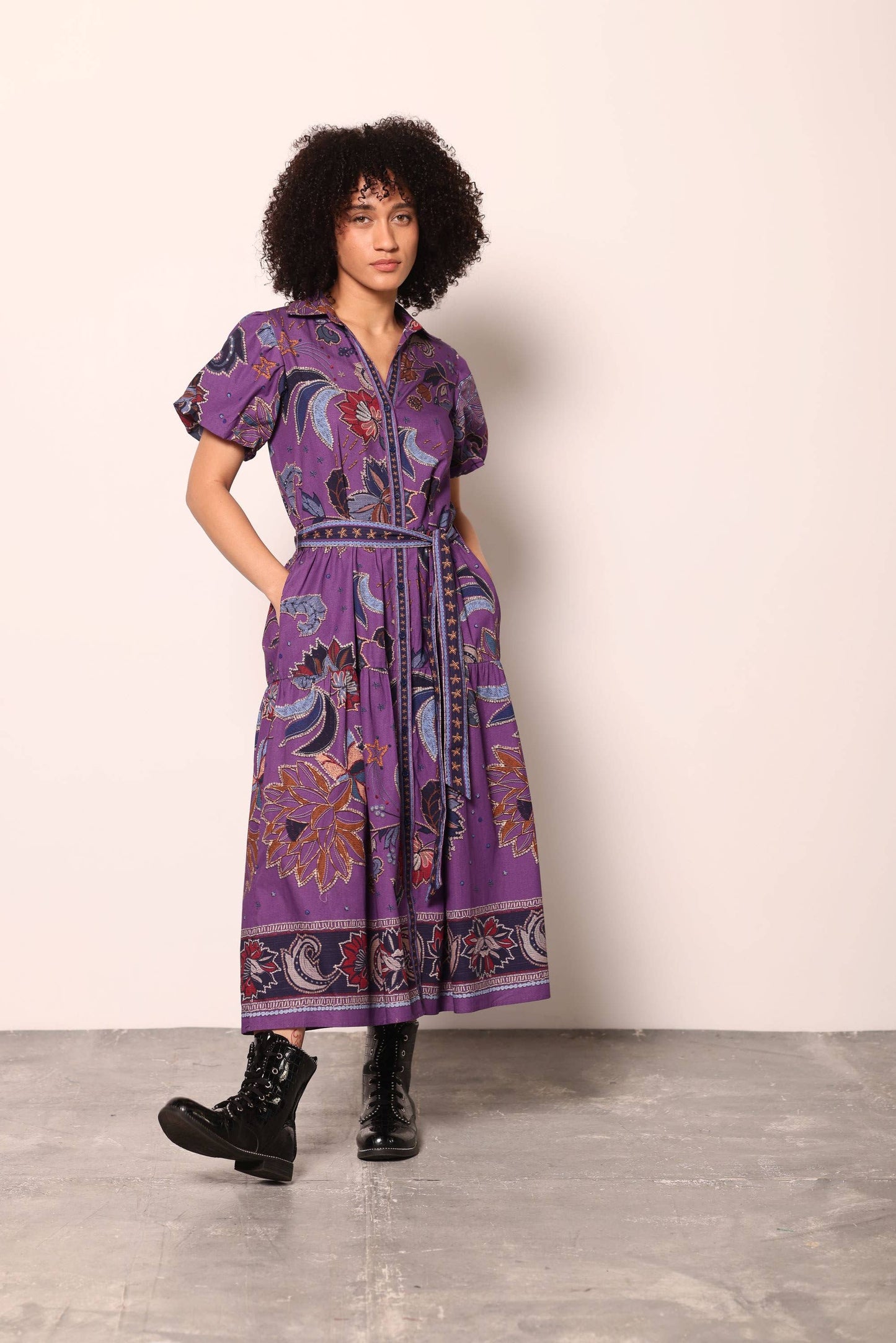 A model with curly black hair stands in a studio wearing a purple short-sleeve midi dress with a colorful paisley and floral print, a matching tie belt, and black combat boots against a light beige wall and gray concrete floor.
